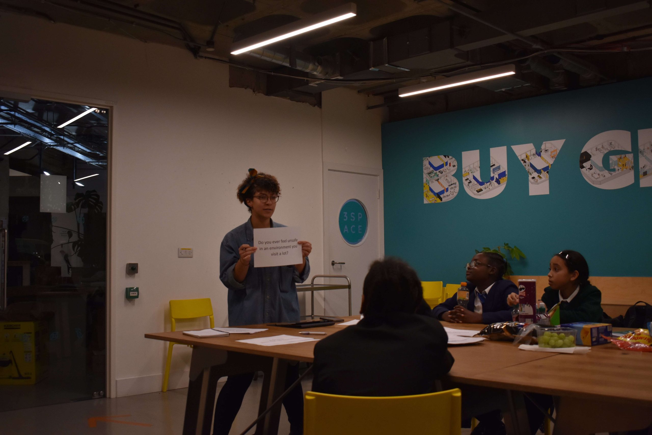 A group of black children sat around a table booadroom style. looking towards a woman holding up a white piece of paper